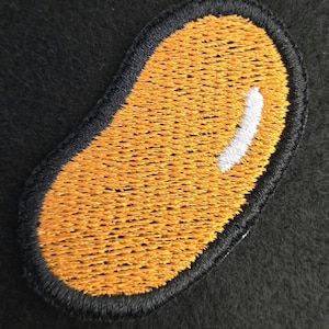 K&ouml;nnte beinhalten: Ein gestickter Aufn&auml;her in Form einer Kidneybohne mit einer leuchtend orangefarbenen F&uuml;llung und einem markanten wei&szlig;en Glanzlichtstich auf der oberen rechten Seite, eingefasst von einem dicken, erhabenen schwarzen Stickrand.