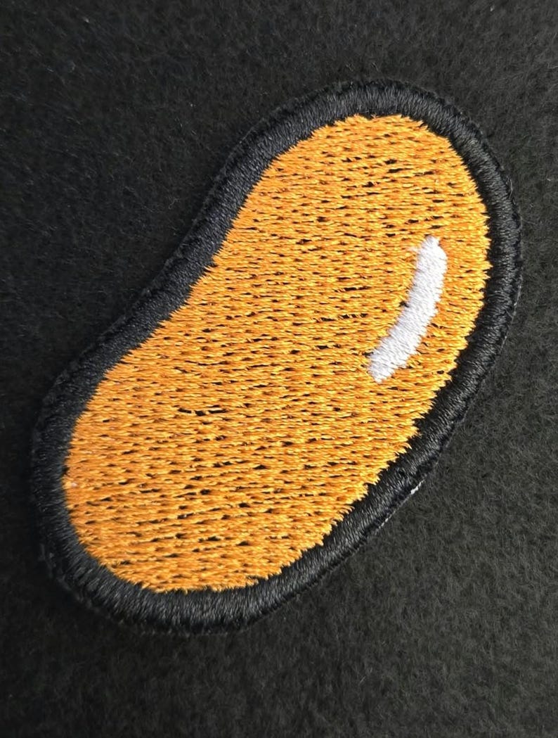 K&ouml;nnte beinhalten: Ein gestickter Aufn&auml;her in Form einer Kidneybohne mit einer leuchtend orangefarbenen F&uuml;llung und einem markanten wei&szlig;en Glanzlichtstich auf der oberen rechten Seite, eingefasst von einem dicken, erhabenen schwarzen Stickrand.