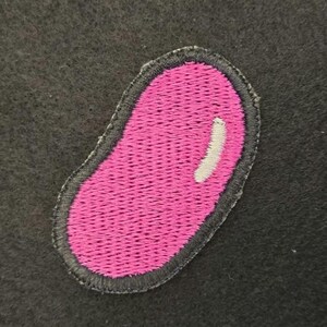 K&ouml;nnte beinhalten: Ein gestickter Aufn&auml;her in Form einer Geleebohne mit leuchtend rosa Mitte und einem kleinen wei&szlig;en Glanzpunkt, eingefasst von einem dunkelgrauen gestickten Rand auf einem dunkelgrauen, filzartigen Stoffhintergrund.