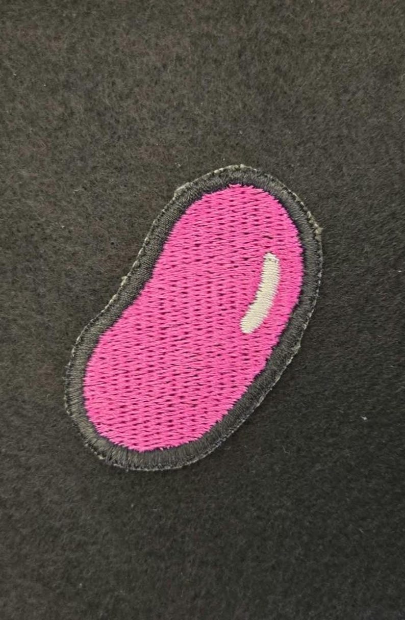 K&ouml;nnte beinhalten: Ein gestickter Aufn&auml;her in Form einer Geleebohne mit leuchtend rosa Mitte und einem kleinen wei&szlig;en Glanzpunkt, eingefasst von einem dunkelgrauen gestickten Rand auf einem dunkelgrauen, filzartigen Stoffhintergrund.