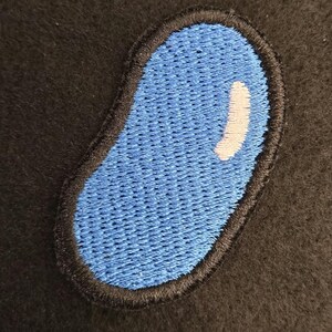 K&ouml;nnte beinhalten: Ein bohnenf&ouml;rmiger Stickaufn&auml;her mit einer leuchtend blauen Mitte und einem kleinen, geschwungenen wei&szlig;en Glanzlicht-Detail. Die gesamte Form ist mit einem dicken, dunkelschwarzen gestickten Rand umrandet, der einen Kontrast bildet.
