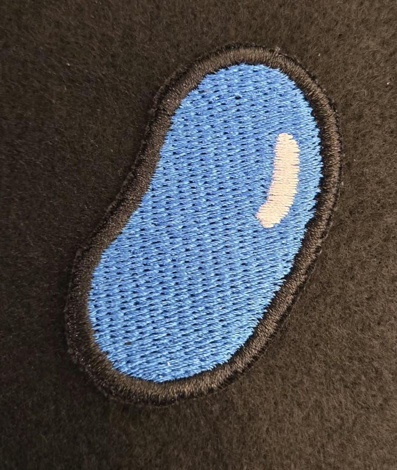 K&ouml;nnte beinhalten: Ein bohnenf&ouml;rmiger Stickaufn&auml;her mit einer leuchtend blauen Mitte und einem kleinen, geschwungenen wei&szlig;en Glanzlicht-Detail. Die gesamte Form ist mit einem dicken, dunkelschwarzen gestickten Rand umrandet, der einen Kontrast bildet.