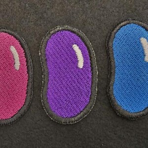 K&ouml;nnte beinhalten: Ein Set aus sieben gestickten Aufn&auml;hern in Form von Geleebohnen, die jeweils eine eigene Volltonfarbe wie Rot, Gelb, Pink, Lila, Blau, Gr&uuml;n und Orange aufweisen, mit einem kleinen, wei&szlig;en, geschwungenen Glanzlicht auf der oberen rechten Seite.