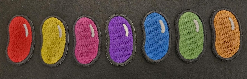 K&ouml;nnte beinhalten: Ein Set aus sieben gestickten Aufn&auml;hern in Form von Geleebohnen, die jeweils eine eigene Volltonfarbe wie Rot, Gelb, Pink, Lila, Blau, Gr&uuml;n und Orange aufweisen, mit einem kleinen, wei&szlig;en, geschwungenen Glanzlicht auf der oberen rechten Seite.