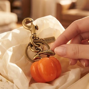 May include: A brass-toned metal keyring featuring a glossy, bright orange pumpkin charm. The keyring includes a lobster claw clasp, a circular ring, and a small rectangular metal tag engraved with the word COACH, all resting in tissue paper.