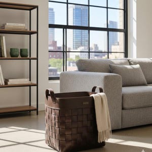May include: A brown woven leather storage basket with two handles, holding a cream-coloured blanket. The basket is in front of a grey sofa and a large window with a city view. A black metal and wood shelf is to the left.