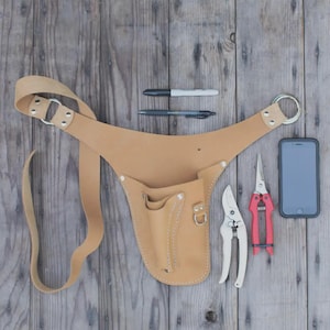 Op de afbeelding: Een beige leren tuingereedschapsriem met zakken, een lange riem en metalen ringen. Twee pennen, een smartphone en twee snoeischaren zijn ook zichtbaar. Eén snoeischaar heeft rode handvatten.
