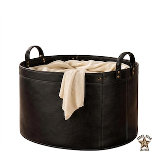 May include: A round black leather storage basket with two sturdy handles attached by brass-coloured rivets. The basket features visible vertical stitching along the sides and contains a soft, folded cream-coloured knit textile inside.