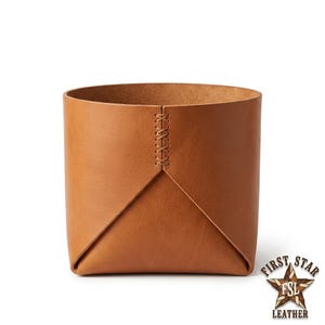 Op de afbeelding: Een beige lederen opbergbak met een cilindrische vorm en een verticale naad verbonden door een kruissteekpatroon. Het leer heeft een gladde, matte afwerking en een consistente lichtbruine kleur over het gehele buitenoppervlak van het object.