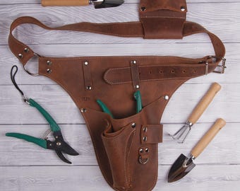 May include: A brown leather gardening tool belt with multiple pockets and a belt. The belt is surrounded by a trowel, a hand rake, a small shovel, and green handled pruning shears. The tools have wooden handles.