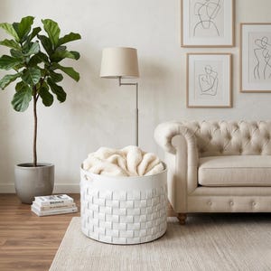 May include: A white woven storage basket filled with a cream-colored blanket sits on a light-colored rug. A floor lamp with a beige lampshade and a beige sofa are in the background. A potted plant and framed art pieces complete the room's decor.