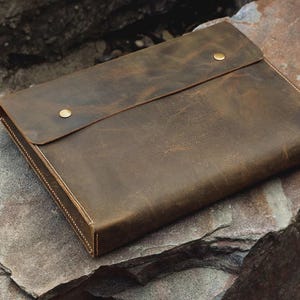 May include: A brown leather portfolio with a flap closure secured by two brass-colored studs. The portfolio is rectangular and appears to be handcrafted, with visible stitching along the edges. The leather has a distressed look.
