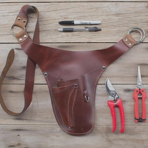 Op de afbeelding: Een bruine leren tuingereedschapsriem met schouderriem, twee pennen en twee rode snoeischaren. De riem heeft meerdere zakken en metalen ringen.