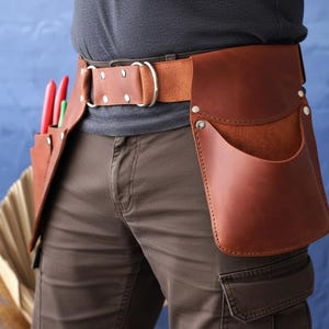 Op de afbeelding: Een bruine leren gereedschapsriem met meerdere zakken en een stevige gesp. De riem heeft een grote zak en verschillende kleinere compartimenten, met rode en groene gereedschappen zichtbaar in een zak. De riem is ontworpen voor het dragen van gereedschap.