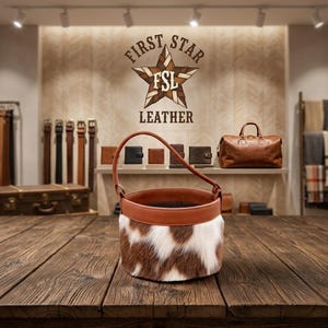 Könnte beinhalten: Eine zylindrische Beuteltasche mit einem Außenmaterial aus braun-weißem Kuhfell, einem glatten braunen Lederrand oben und einem einzelnen passenden Ledergriff, platziert auf einer dunklen Holzoberfläche in einem Lederwarengeschäft.
