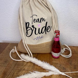 Pode incluir: Uma bolsa bege com cordão com o texto "Team Bride" em escrita preta e um pequeno coração rosa. Uma pequena garrafa de bebida rosa e grama decorativa branca também estão presentes. A bolsa tem uma base de serapilheira.