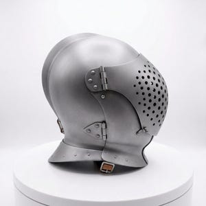May include: A silver-colored medieval helmet with a rounded top and a hinged visor featuring small, evenly spaced holes. The helmet has riveted details and a chin strap, suggesting historical armor.