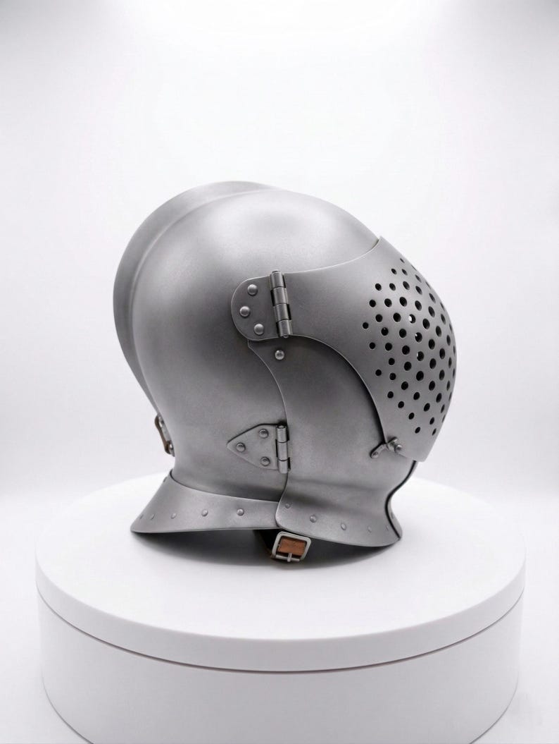 May include: A silver-colored medieval helmet with a rounded top and a hinged visor featuring small, evenly spaced holes. The helmet has riveted details and a chin strap, suggesting historical armor.