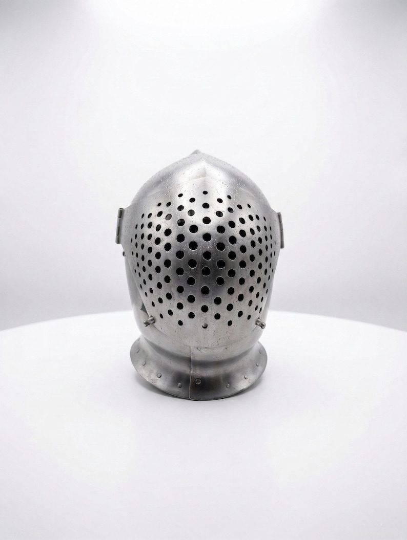 May include: A silver-colored medieval helmet with a rounded top and a faceplate covered in small, evenly spaced holes. The helmet sits on a white surface, showcasing its metallic sheen and historical design.