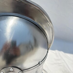 May include: A polished silver-colored metal helmet featuring a rounded dome, a wide brim, and a reinforced band secured with small circular rivets. The surface is smooth and reflective, showing a metallic sheen with visible construction details.