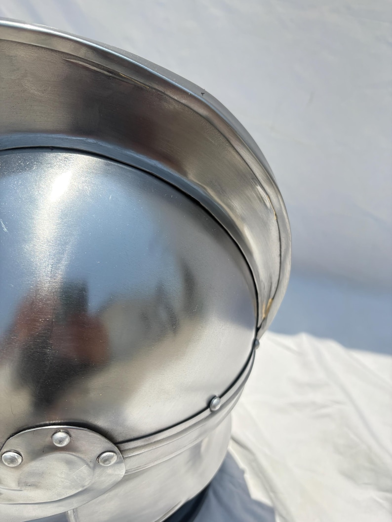 May include: A polished silver-colored metal helmet featuring a rounded dome, a wide brim, and a reinforced band secured with small circular rivets. The surface is smooth and reflective, showing a metallic sheen with visible construction details.