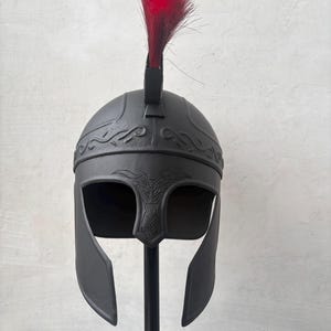 May include: A matte black helmet featuring a wide face opening, a nose guard with etched detailing, and a decorative band across the forehead. A vibrant red crest made of stiff, upright bristles is attached to the top center of the helmet.