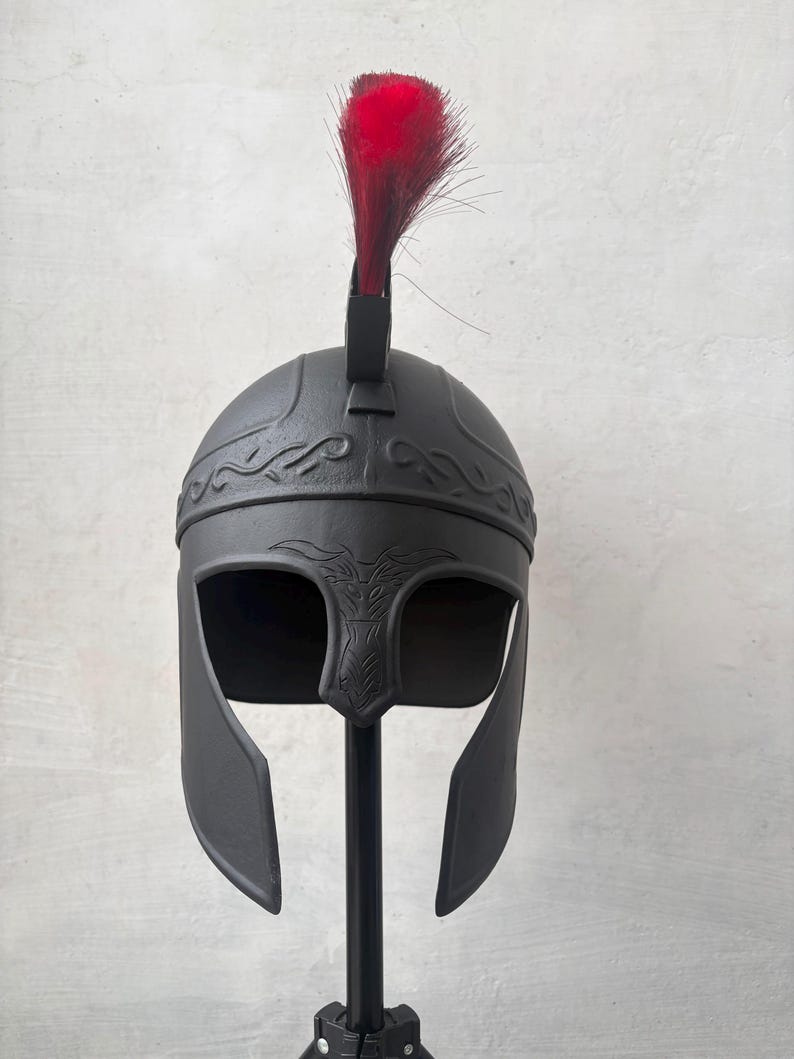 May include: A matte black helmet featuring a wide face opening, a nose guard with etched detailing, and a decorative band across the forehead. A vibrant red crest made of stiff, upright bristles is attached to the top center of the helmet.