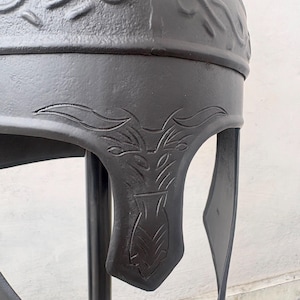 May include: A matte black metal helmet featuring embossed scrollwork patterns along the crown and intricate engraved floral or leaf-like motifs on the cheek guard. The helmet has a smooth, dark finish and a structured design with a curved visor.