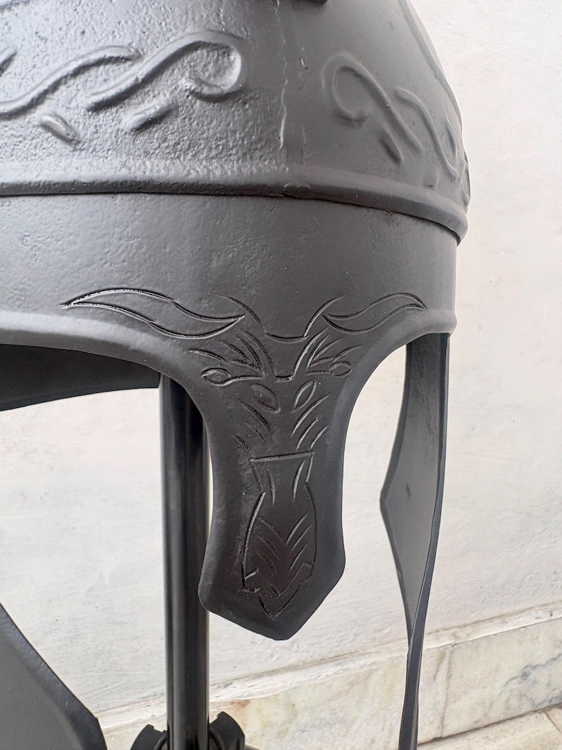 May include: A matte black metal helmet featuring embossed scrollwork patterns along the crown and intricate engraved floral or leaf-like motifs on the cheek guard. The helmet has a smooth, dark finish and a structured design with a curved visor.