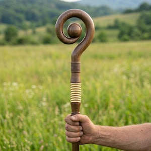 Op de afbeelding: Een houten staf met een donkerbruine afwerking en een opvallende spiraalvormige top met een centrale bol. De schacht bevat een reeks horizontale groeven en een sectie met heldere goudkleurige ringen onder de spiraalvormige kop.