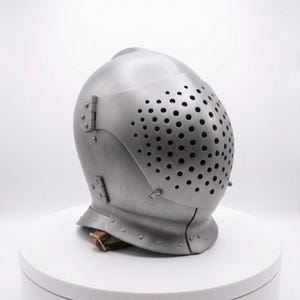 May include: A silver-colored medieval helmet with a perforated visor. The helmet has a rounded top and is secured with hinges and a leather strap. The visor is covered in small, evenly spaced holes.