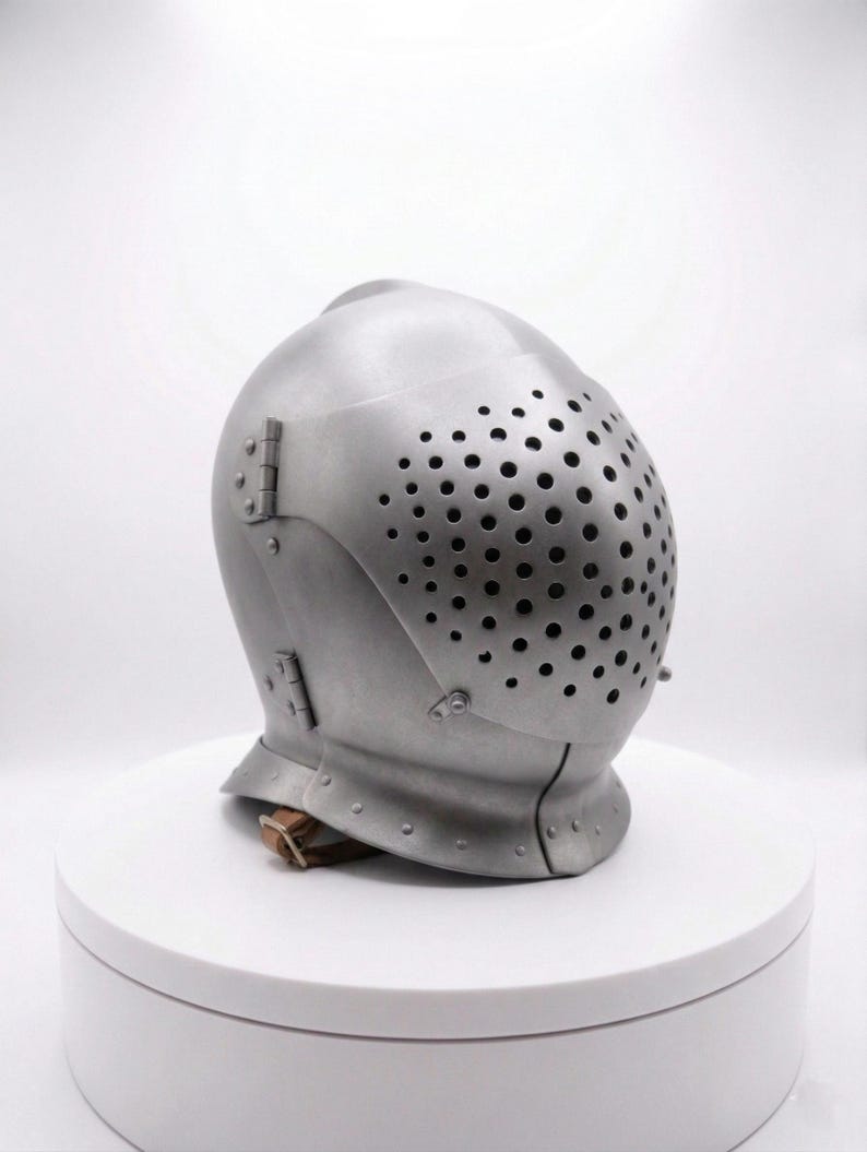 May include: A silver-colored medieval helmet with a perforated visor. The helmet has a rounded top and is secured with hinges and a leather strap. The visor is covered in small, evenly spaced holes.
