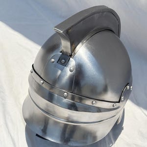 May include: A polished steel helmet featuring a rounded dome, a prominent central crest running from front to back, and a wide, curved brim. The metal surface is secured with visible rivets along the base of the crest and the lower rim.