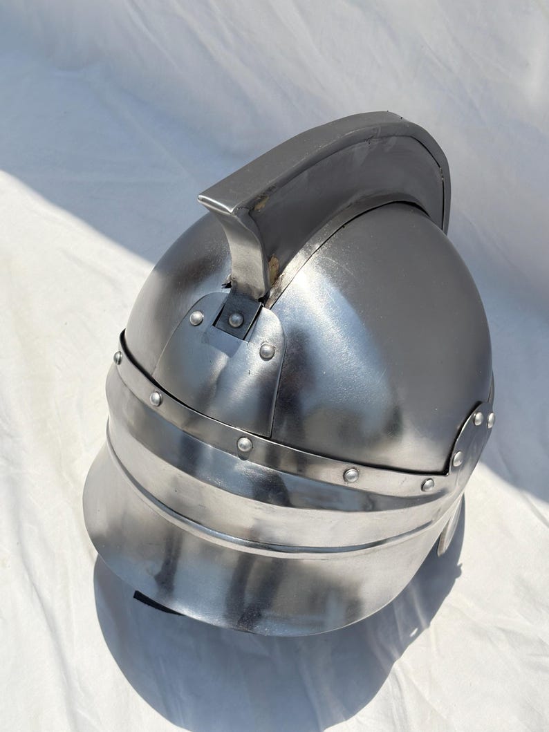 May include: A polished steel helmet featuring a rounded dome, a prominent central crest running from front to back, and a wide, curved brim. The metal surface is secured with visible rivets along the base of the crest and the lower rim.
