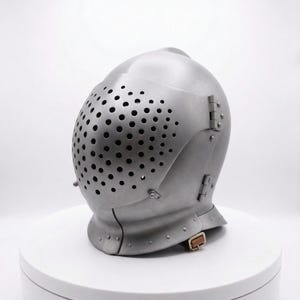 Puede incluir: Un casco medieval plateado con una parte superior redondeada y una placa frontal cubierta de pequeños agujeros espaciados uniformemente. El casco tiene secciones con bisagras y una correa para la barbilla con hebilla. Se exhibe sobre una plataforma circular blanca.