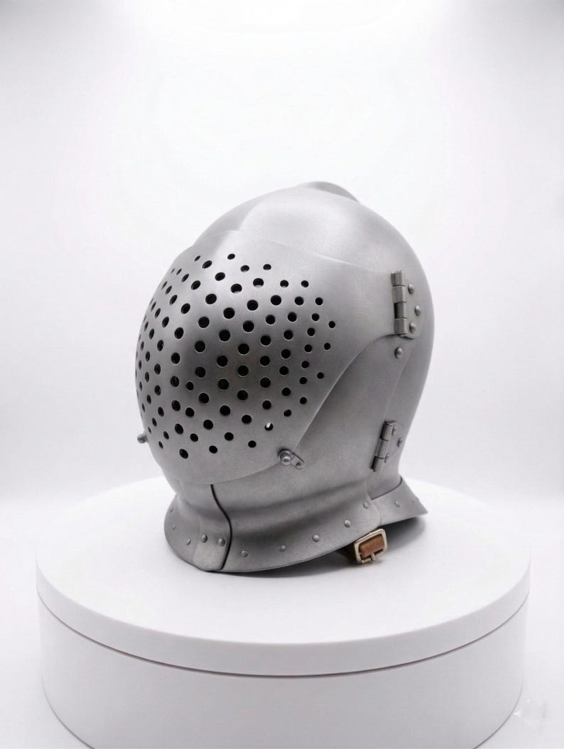 May include: A silver-colored medieval helmet with a rounded top and a faceplate covered in small, evenly spaced holes. The helmet has hinged sections and a chin strap with a buckle. It is displayed on a white, circular platform.
