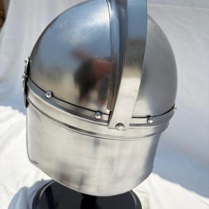 May include: A polished silver-colored metal helmet featuring a rounded dome, a prominent vertical central ridge, and a horizontal band secured with small rivets. The helmet is mounted on a circular black base, showcasing a smooth, reflective surface.