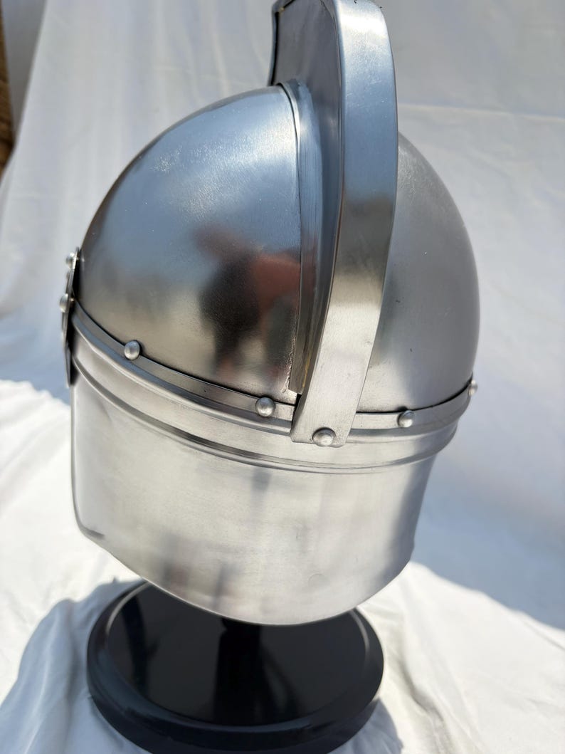 May include: A polished silver-colored metal helmet featuring a rounded dome, a prominent vertical central ridge, and a horizontal band secured with small rivets. The helmet is mounted on a circular black base, showcasing a smooth, reflective surface.