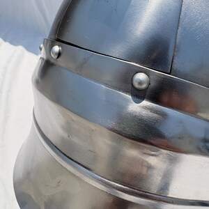 May include: A polished silver-colored metal helmet featuring a rounded dome construction with a reinforced horizontal band. The surface is accented with multiple visible rivets securing the overlapping metal plates together in a structured design.
