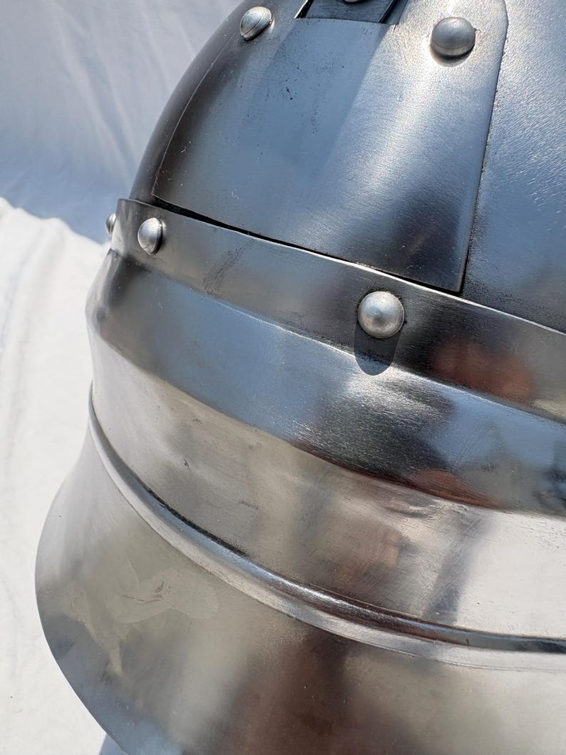 May include: A polished silver-colored metal helmet featuring a rounded dome construction with a reinforced horizontal band. The surface is accented with multiple visible rivets securing the overlapping metal plates together in a structured design.