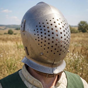 May include: A silver-colored medieval helmet with numerous small ventilation holes covering the face area. The helmet has a rounded top and a protective neck guard. The person is wearing a green vest over a quilted undergarment, suggesting historical reenactment or costume.