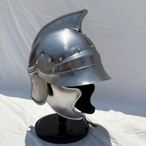 May include: A polished silver-colored metal helmet featuring a prominent central crest, a curved visor, and side cheek guards. The helmet is constructed with visible rivets along the brow and side panels, presenting a smooth, reflective surface.