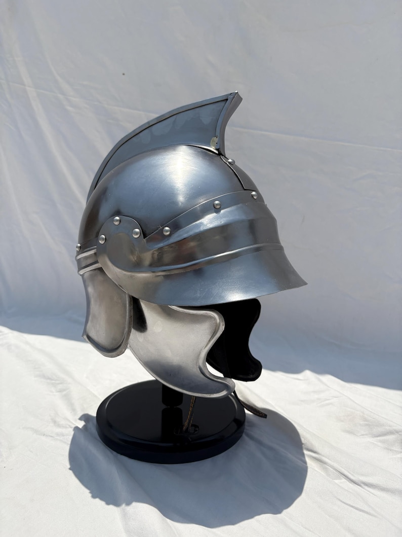 May include: A polished silver-colored metal helmet featuring a prominent central crest, a curved visor, and side cheek guards. The helmet is constructed with visible rivets along the brow and side panels, presenting a smooth, reflective surface.