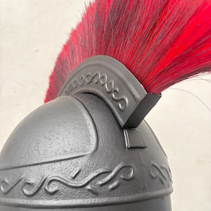 May include: A matte black helmet featuring a prominent crest with long, vibrant red bristles. The helmet is decorated with embossed scrollwork patterns along the crown and the base of the crest, showcasing a textured, dark finish throughout.