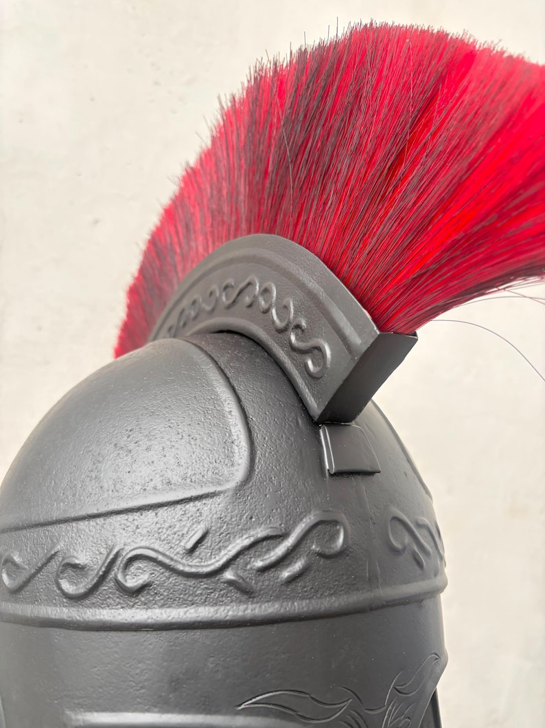 May include: A matte black helmet featuring a prominent crest with long, vibrant red bristles. The helmet is decorated with embossed scrollwork patterns along the crown and the base of the crest, showcasing a textured, dark finish throughout.