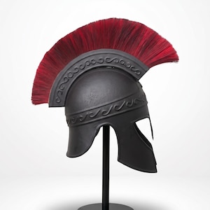 May include: A matte black Roman-style helmet featuring a prominent crest of long, vibrant red bristles. The helmet is adorned with an embossed scrollwork pattern along the brow and the base of the crest, mounted on a simple black display stand.