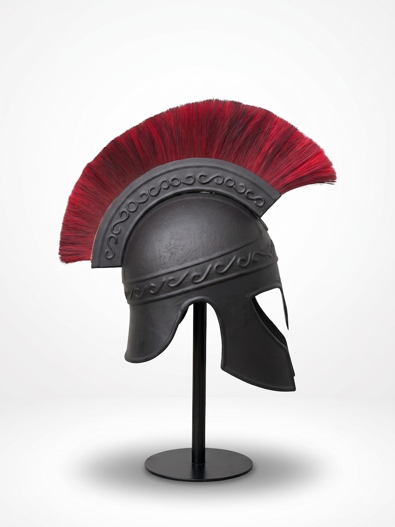 May include: A matte black Roman-style helmet featuring a prominent crest of long, vibrant red bristles. The helmet is adorned with an embossed scrollwork pattern along the brow and the base of the crest, mounted on a simple black display stand.