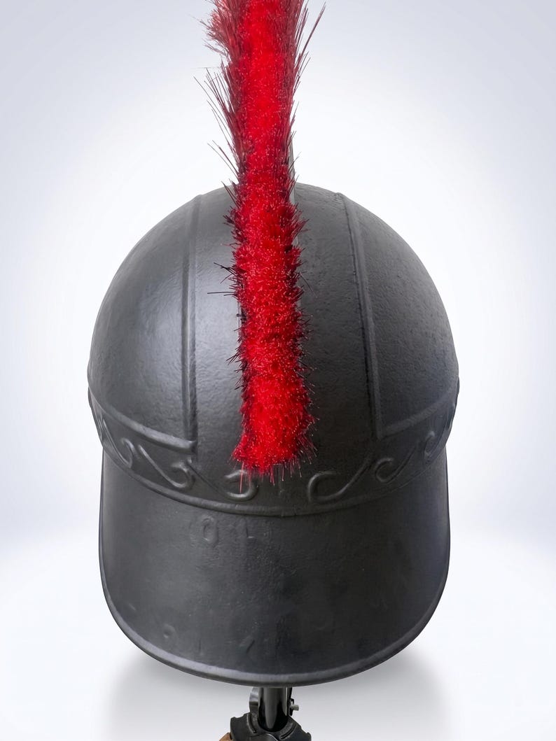 May include: A matte black helmet featuring a prominent, upright red crest made of stiff, fibrous material. The helmet has a smooth, rounded dome with a raised horizontal band across the front, decorated with a subtle, embossed wave-like pattern.