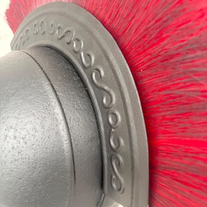 May include: A matte black helmet featuring a prominent crest adorned with a raised, repeating S-shaped scroll pattern. A dense, vibrant red brush-like plume extends vertically from the top of the curved crest, creating a striking contrast.