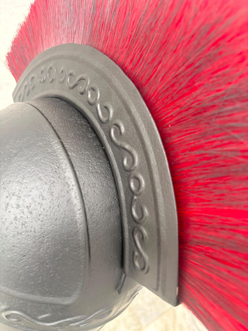 May include: A matte black helmet featuring a prominent crest adorned with a raised, repeating S-shaped scroll pattern. A dense, vibrant red brush-like plume extends vertically from the top of the curved crest, creating a striking contrast.